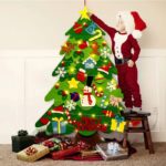 Ein als Weihnachtsmann verkleidetes Kind steht neben einem Weihnachtsbaum aus Filz - 26 Deko-Teile + Lichterkette - 95cm, der mit Filzstift verziert ist.