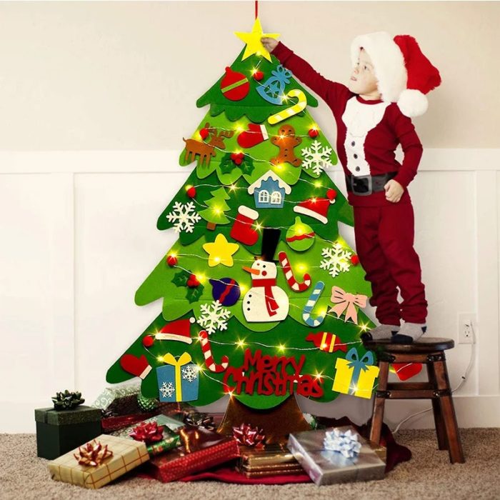 Ein als Weihnachtsmann verkleidetes Kind steht neben einem Weihnachtsbaum aus Filz - 26 Deko-Teile + Lichterkette - 95cm, der mit Filzstift verziert ist.