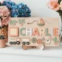 Ein hölzernes Namenspuzzle mit der Aufschrift "CHARLIE" besteht aus abnehmbaren Buchstaben und Formen, darunter ein Dinosaurier, ein Boot, ein Zug, ein Kaktus und Zahnrädern, und wird auf einem Ständer mit Blumen im Hintergrund präsentiert.